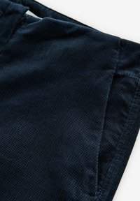 Material de catifea de un albastru închis, cu textură verticală cu caneluri, care arată o parte din talia și detaliul buzunarului unui articol de vestimentație.