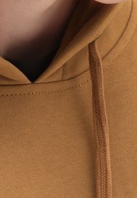 Sudadera marrón con cordón, que presenta una textura suave, puños acanalados y un bolsillo tipo canguro. El tejido parece liso y ligero.