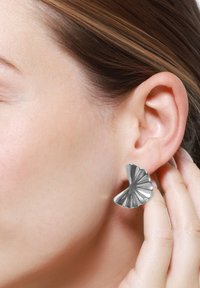 Sølvfargede metaløredobber i en vifteform med teksturerte, ribbede detaljer. Båret på loben, som viser et strømlinjeformet og moderne design.