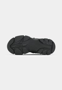 Semelle de chaussure en caoutchouc noir avec un motif de bande de roulement prononcé et le logo "LACOSTE" embossé au centre.