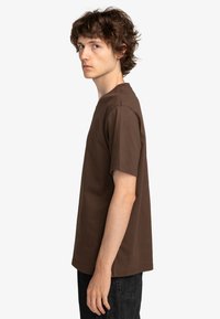 Brown cotton t-shirt with short sleeves, crew neck, straight cut, and minimal stitching details. Worn with dark jeans.