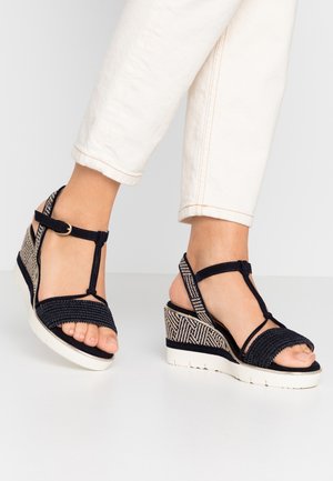 Feet wearing black woven wedge sandals with ankle straps and white soles, paired with rolled-up white pants.