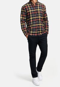 Homme portant une chemise à carreaux à manches longues boutonnée en noir, jaune et rouge, un pantalon noir et des baskets blanches, debout les mains dans les poches.