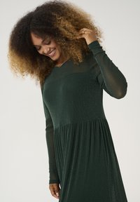 Knee-length dark green dress with long sleeves and a sheer bodice. Features a gathered skirt and a subtle glitter texture throughout.