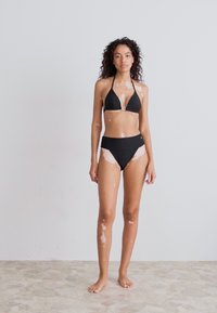 Black bikini set featuring a triangular top and high-waisted bottom. Model stands barefoot, displaying skin with light patches against a neutral background.