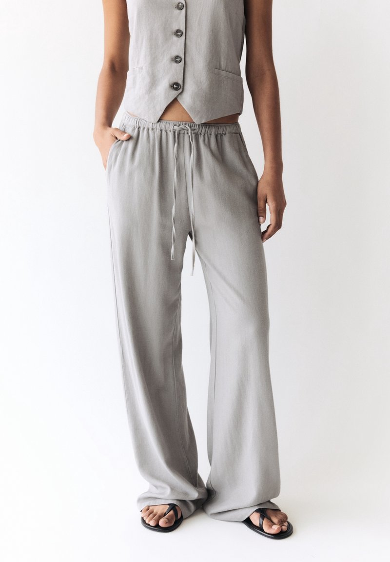 Femme portant un pantalon large gris clair à cordon de serrage, un gilet sans manches boutonné et des tongs noires sur fond blanc.
