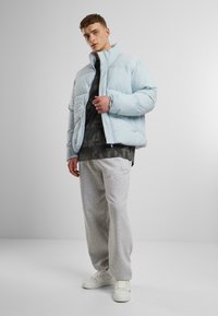 Ljusblå puffjacka med hög krage, kviltat mönster, bärs över en mörk kamouflage tröja och grå sweatpants, kompletterad med vita sneakers.