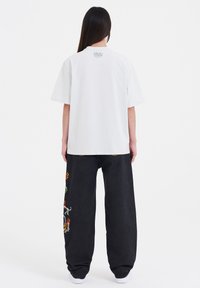 Witte t-shirt met korte mouwen en een ronde halslijn, voorzien van een logo op de achterkant, gecombineerd met zwarte jeans met kleurrijke draakborduursels.