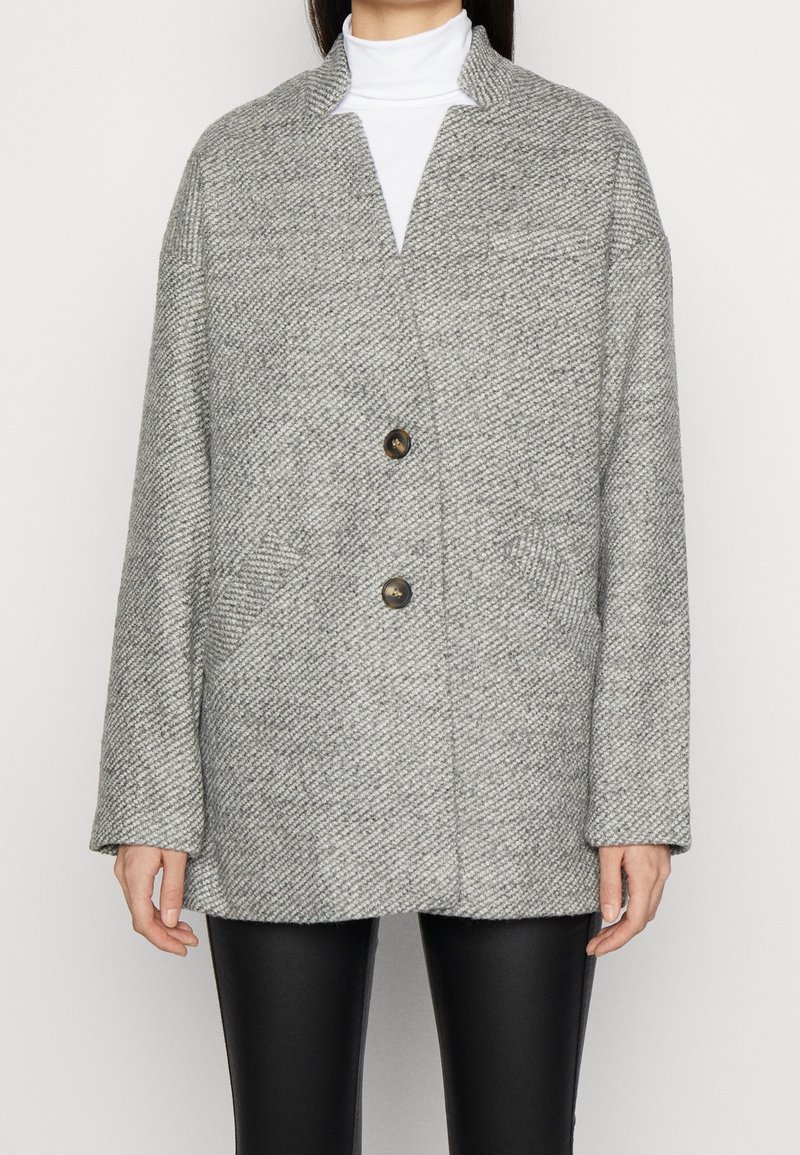 Grauer strukturierter Mantel mit einem Kragen, zwei Fronttaschen und zwei Knöpfen. Lange Ärmel und lockere Passform, über einem weißen Rollkragenpullover getragen.