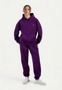 Fioletowa bluza z kapturem i dopasowane spodnie dresowe wykonane z miękkiej tkaniny. Bluza wyposażona w przednią kieszeń i ściągacze przy rękawach; strój zestawiony z białymi trampkami.