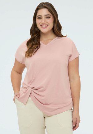 Smilende kvinde med langt, bølget hår iført en lyserød kortærmet top med knude i siden og beige bukser foran en ensfarvet baggrund.