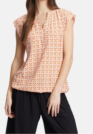 Femme portant un haut sans manches à motifs géométriques orange et blanc avec un col en V, assorti à un pantalon noir ample à plis.