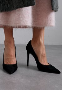 Černé semišové lodičky na vysokém podpatku s špičatou špičkou a tenkým stiletto podpatkem. Boty mají hladký povrch a minimalistický design.
