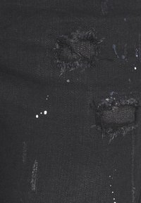 Černá denimová tkanina s popraskanými místy, se třepenými okraji a bílými skvrnami od barvy, která vytváří strukturovanou, městskou estetiku.