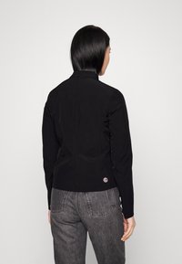 Černá bunda s vysokým límcem, se slim fit střihem a malým kulatým logem na zádech, nošená s šedými džínovými kalhotami.