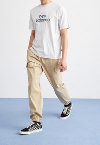 T-shirt gris clair avec le texte "new balance", pantalon cargo beige et baskets à carreaux noir et blanc, marchant sur un sol bleu.