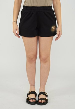 Vrouw draagt zwarte shorts met elastische taille en gouden EA7-logo en zwarte sandalen met gouden gespen, staand op een effen achtergrond.