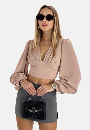 Blouse croppée rose pâle avec des manches volumineuses, assortie à une mini-jupe grise. Les accessoires comprennent un petit sac à main noir texturé et une montre argentée.