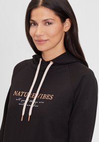 Sudadera negra con el texto "NATURE VIBES" en oro rosado metálico, con cordones color crema y una textura suave. Bordado blanco debajo del texto.