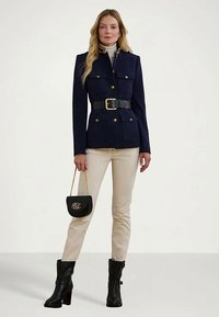 Γυναίκα φορώντας ναυτικό μπουφάν με ζώνη, μπεζ παντελόνι, μαύρες τακούνες μπότες και κρατώντας μια μαύρη τσάντα με χρυσή αλυσίδα και αρχικά.