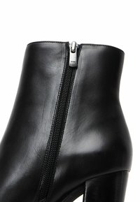 Bottine noire en cuir avec fermeture éclair latérale et talon bloc, texture lisse, gros plan sur la partie supérieure et la zone de la fermeture éclair.