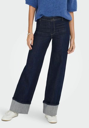 Jean en denim foncé à jambes larges avec des poignets effilochés gris. Associé à un haut en tricot bleu ample et des baskets blanches. Texture lisse, design décontracté.