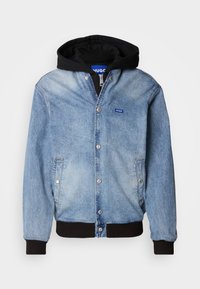 Denim jacket featuring light blue fabric, snap-button closure, side pockets, and a black hood. Branding displayed on the chest.