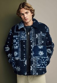 Marineblauwe fleecejas met een patchworkontwerp en bloemmotieven, drukknoopsluiting en een klassieke kraag. Zachte, gestructureerde stof.