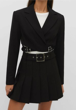 Vrouw met een zwart cropped blazer met een dubbele gespsluiting over een wit topje, gecombineerd met een zwarte, geplooide rok met hoge taille.