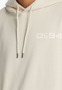 Béžová mikina s měkkou strukturou, vybavená kapucí na šňůrku a logem "DC94" v mírně tmavším odstínu.