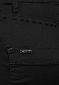 Jeans de mezclilla negra con una textura suave, con costuras prominentes, una pequeña placa de logo plateada y un bolsillo frontal con un remache de metal.