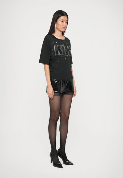 Black graphic t-shirt with rhinestone lettering, paired with glossy black shorts and sheer black tights. Model wears pointed black heels.