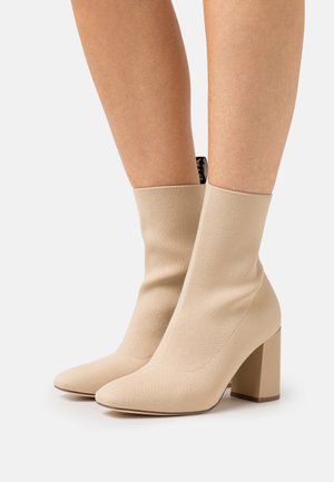Botas de tobillo de punto beige con punta afilada, caña alta elástica y tacón cuadrado. El material texturizado proporciona un ajuste ceñido y un diseño moderno.