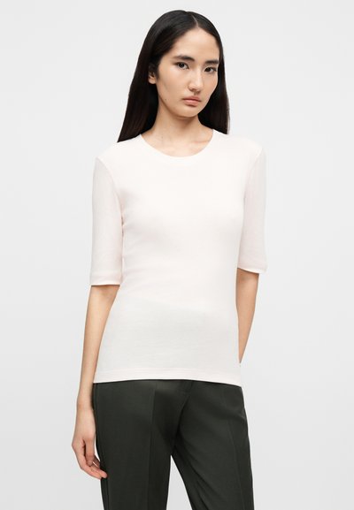 Γυναίκα με μακριά σκούρα μαλλιά που φοράει ανοιχτό ροζ κοντομάνικο τοπ και σκούρο γκρι παντελόνι, στέκεται μπροστά σε απλό λευκό φόντο.