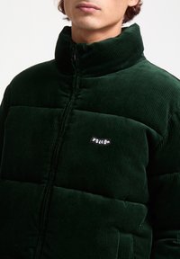 Green corduroy puffer jacket with a high collar, featuring horizontal quilted sections and a small black "Volcom" logo on the chest.