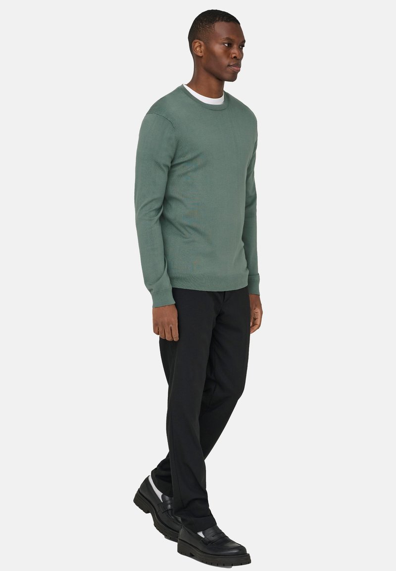 Homme portant un pull vert, un pantalon noir et des mocassins noirs, marchant devant un fond clair uni.