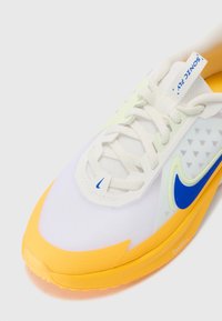 Hvit sneaker med gul såle, blå detaljer og teksturert overdel. Har dekorativt trekantmønster og snøringslukking for en trygg passform.