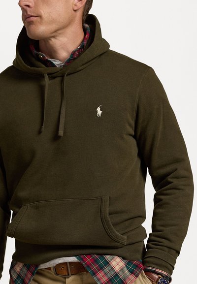 Haut à capuche vert olive avec une poche kangourou, un cordon de serrage pour la capuche et un petit logo brodé. Superposé avec une chemise à carreaux en dessous.