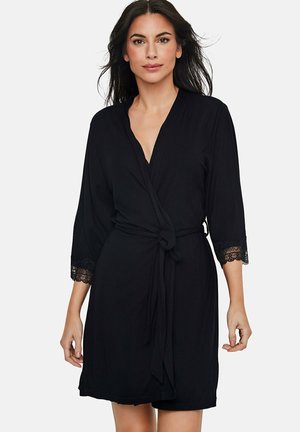 Femme portant une robe portefeuille noire longueur genou avec bordure en dentelle aux manches 3/4, attachée à la taille avec une ceinture en tissu assorti.