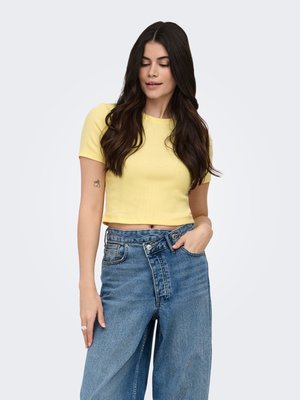 Woman with long dark hair wearing a yellow cropped t-shirt and high-waisted loose blue jeans with an asymmetrical button fly.