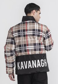 Nafukovacia bunda s hnedo-červeným a čiernym károvaným vzorom, čierna spodná časť s bielym textom "KAVANAGH", vysoký golier a prešívaná textúra.