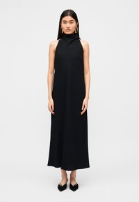 Μαύρο αμάνικο φόρεμα με ψηλό λαιμό, ρέον σχέδιο και διακριτή υφή· συνοδεύεται από μαύρες μπαλαρίνες με μυτερές άκρες.