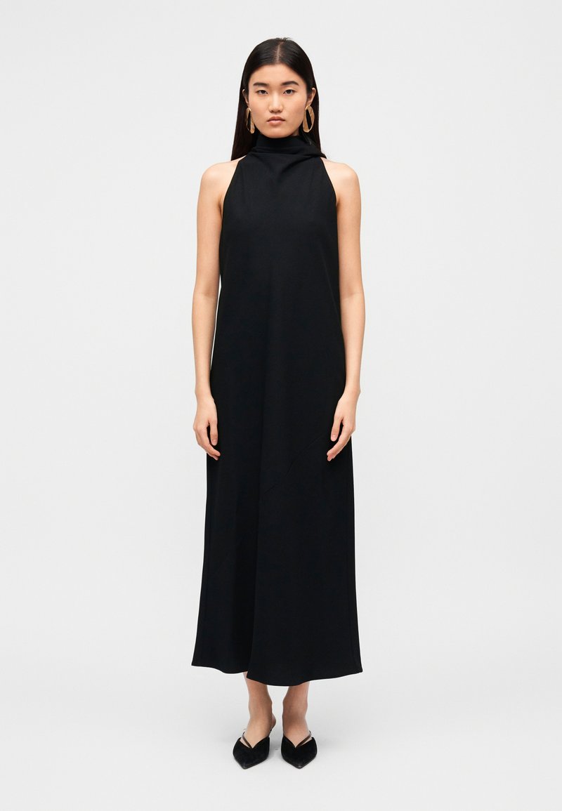 Μαύρο αμάνικο φόρεμα με ψηλό λαιμό, ρέον σχέδιο και διακριτή υφή· συνοδεύεται από μαύρες μπαλαρίνες με μυτερές άκρες.