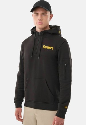 Giovane uomo con felpa nera dei Pittsburgh Steelers con zip frontale e cappello da baseball nero su sfondo uniforme.