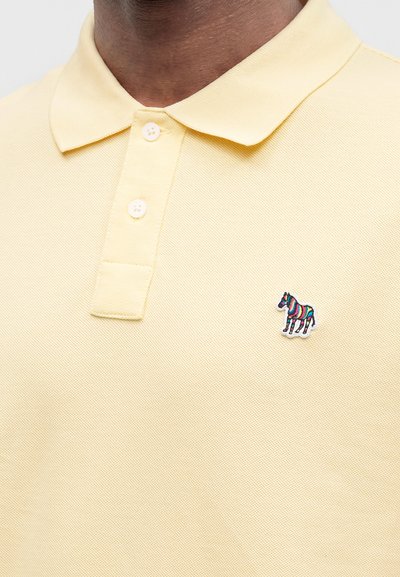 Gros plan d’une personne portant un polo jaune pâle à deux boutons avec un petit logo zèbre coloré sur la poitrine.