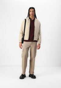 Veste bomber beige sur un pull en maille bordeaux, pantalon cargo beige et chaussures à plateforme noires. Comprend un sac crossbody noir et une coiffure en nattes.
