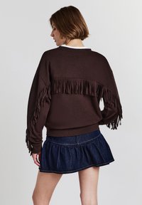 Felpa marrone con dettagli frangiati sulle maniche, abbinata a una gonna a campana in jeans scuri. Tessuto morbido e design a vestibilità rilassata.