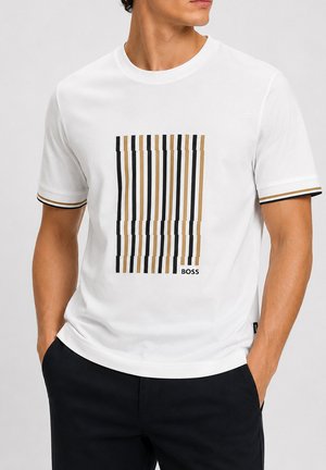 Uomo che indossa una T-shirt bianca a maniche corte con strisce verticali nere e beige e la scritta "BOSS", abbinata a pantaloni neri.