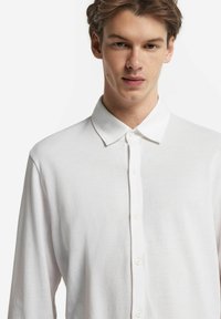 Camicia bianca a maniche lunghe realizzata in tessuto leggero, con colletto classico, chiusura con bottoni e texture minimale.