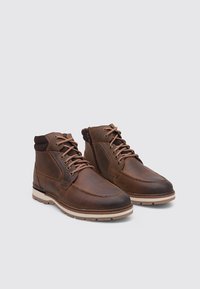 Botines de cuero marrón con punta redondeada, acabado texturizado y suela clara en contraste. Cuentan con cordones marrones y detalle de cremallera lateral.